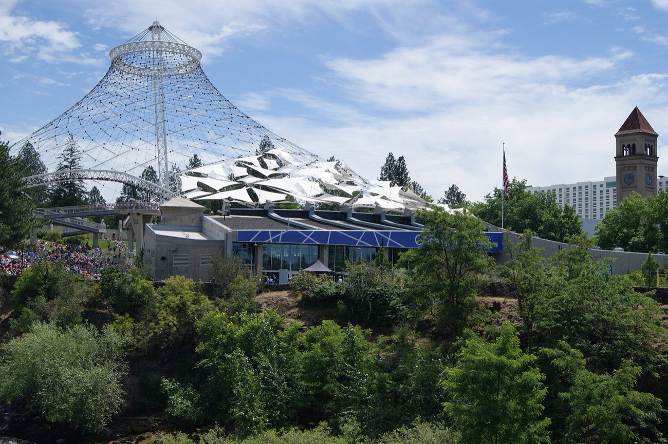 A large crowd on the hillside by the Pavilion in Riverfront Park, its net structure overhead, with the Great Northern clock tower in the distance under a summer sky.