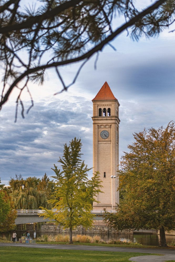 The Great Northern clock tower in Riverfront Park, framed by trees, with the Howard Street bridge and the Spokane River behind it.