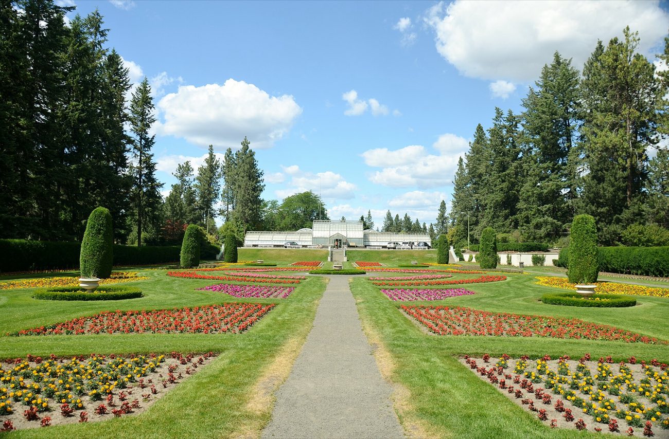Duncan Garden at Manito Park in full summer bloom: formal flower beds, clipped hedges, a gravel path leading to the glass conservatory, ponderosa pines behind.